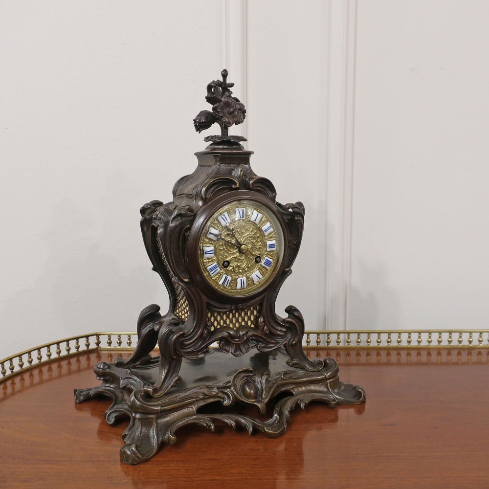A patinated dark bronze French Rococo style mantel clock featuring scrolling acanthus motifs, a floral finial, and blue Roman numerals on individual enamel cartouches.