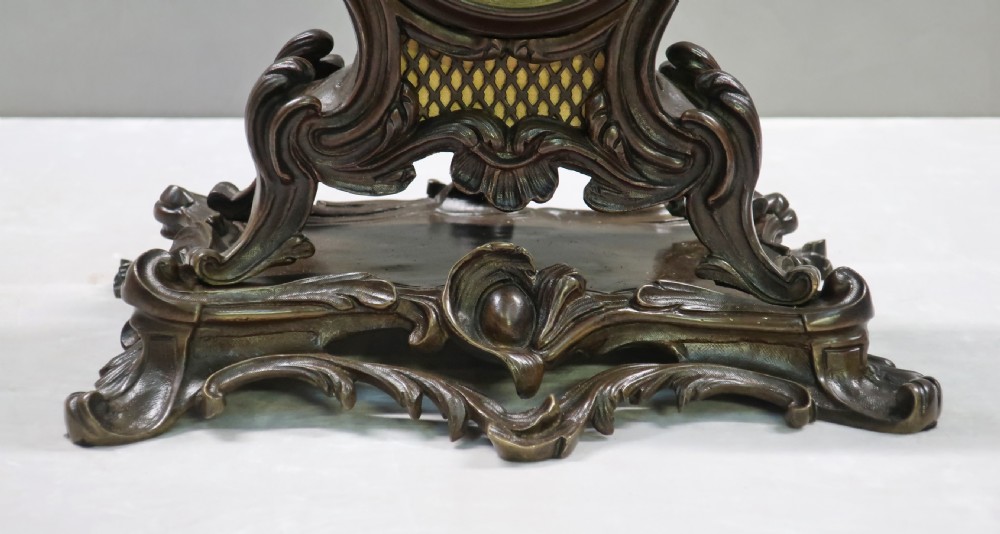 A patinated dark bronze French Rococo style mantel clock featuring scrolling acanthus motifs, a floral finial, and blue Roman numerals on individual enamel cartouches. View IV
