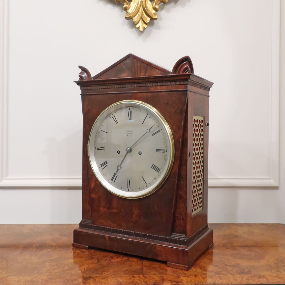 A Georgian flame mahogany bracket clock featuring a circular white dial with Roman numerals, a pointed architectural pediment, and brass side frets.