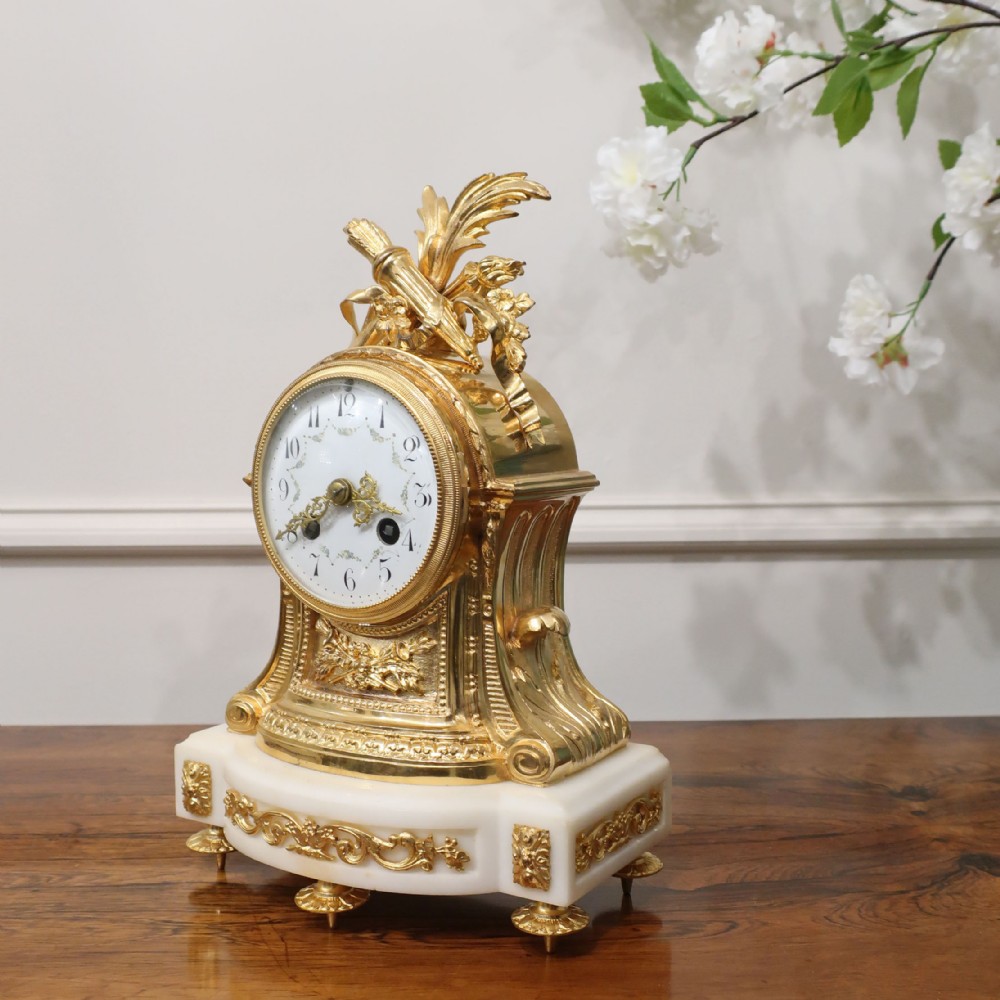 An ornate French gilt bronze and white marble mantel clock featuring a white enamel dial topped with leaf motifs.
