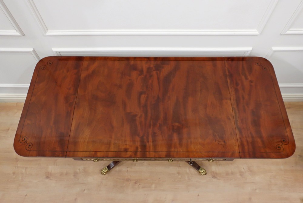 A Regency mahogany sofa table featuring two drop leaves, two front drawers with brass knobs, decorative brass mounts, and splayed legs resting on brass lion paw castors. View III