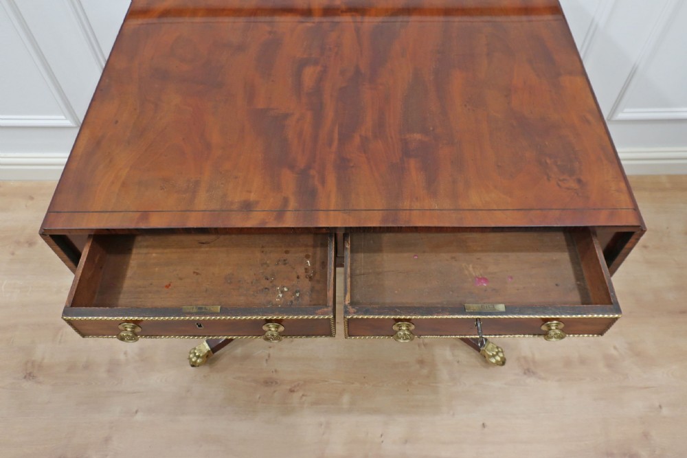 A Regency mahogany sofa table featuring two drop leaves, two front drawers with brass knobs, decorative brass mounts, and splayed legs resting on brass lion paw castors. View X