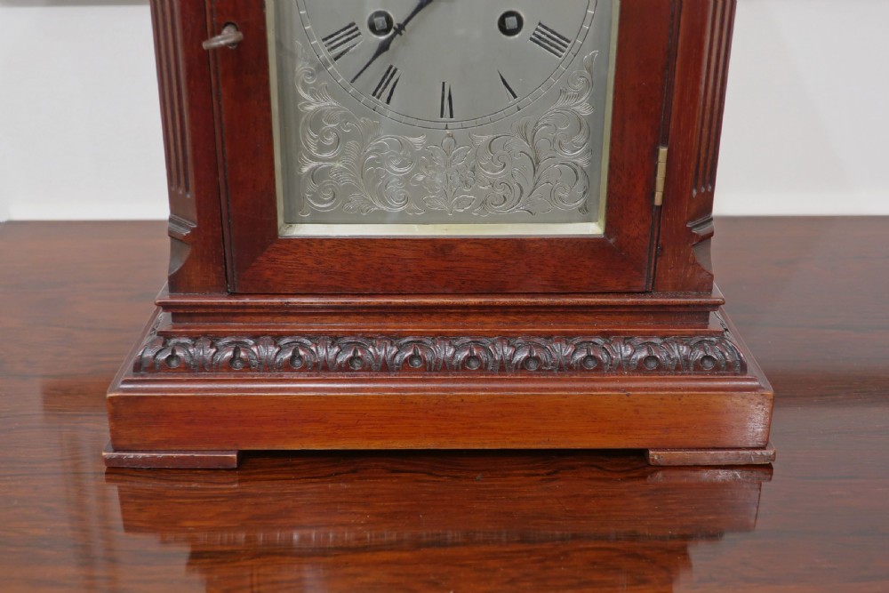 A dark mahogany mantel clock featuring a carved top pediment, fluted side pilasters, and a silvered metal dial with Roman numerals behind a hinged glass door. View V