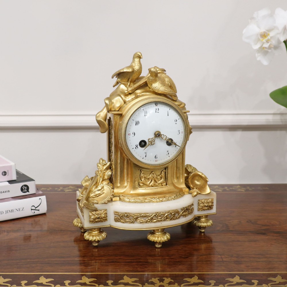 A French Louis XVI style gilt bronze and white marble mantel clock featuring a pair of sculpted doves atop a circular enamel dial flanked by fluted pillars.