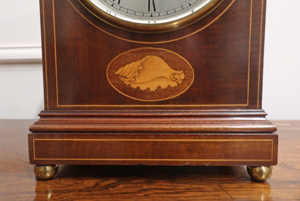 An Edwardian arched mahogany mantel clock featuring line inlay, a central shell marquetry motif, brass ball feet, and a silvered dial with Roman numerals. View IV