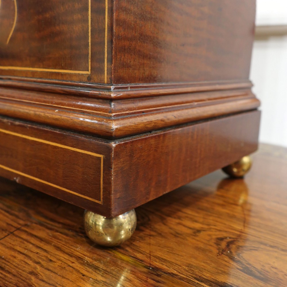 An Edwardian arched mahogany mantel clock featuring line inlay, a central shell marquetry motif, brass ball feet, and a silvered dial with Roman numerals. View V