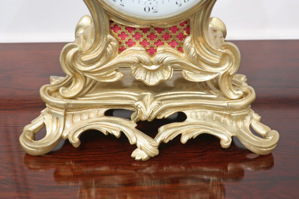 A French Louis XV style gilt brass mantel clock topped with a seated cherub figurine and featuring an ornate Rococo scroll base surrounding a white enamel dial. View V