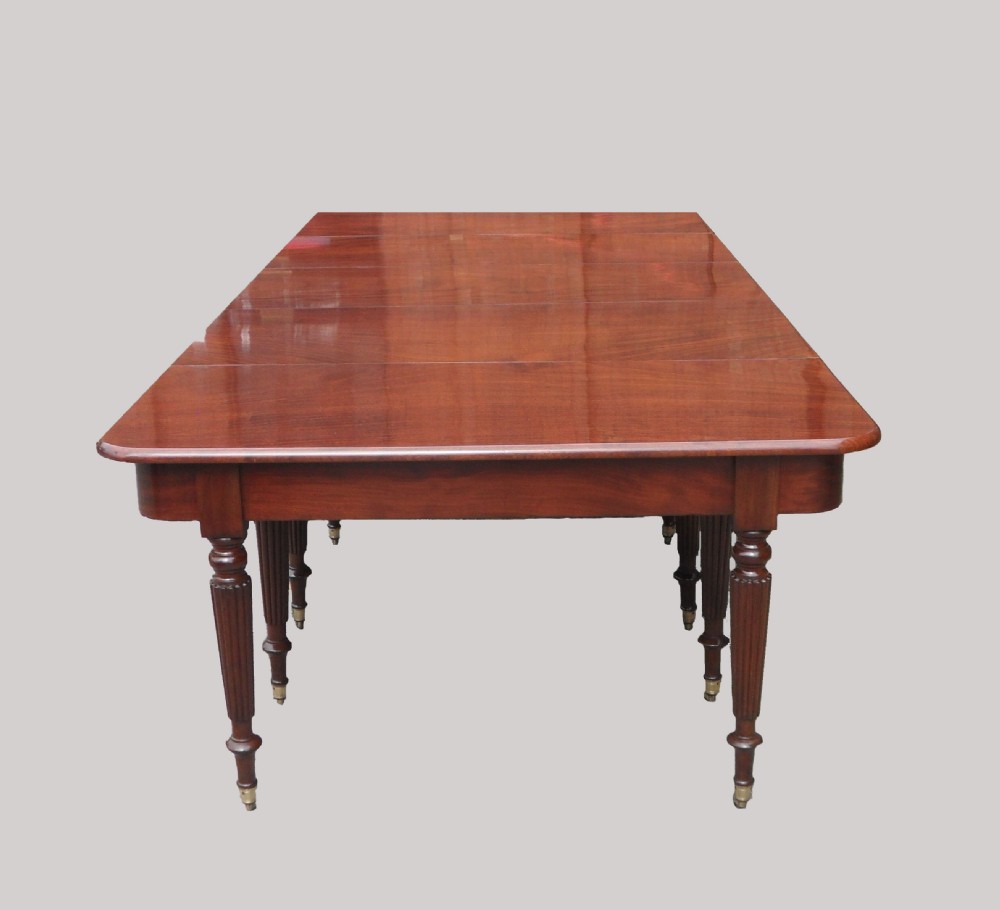 A long Georgian mahogany extending dining table featuring a polished rectangular top with rounded corners supported by reeded legs on brass castors. View II
