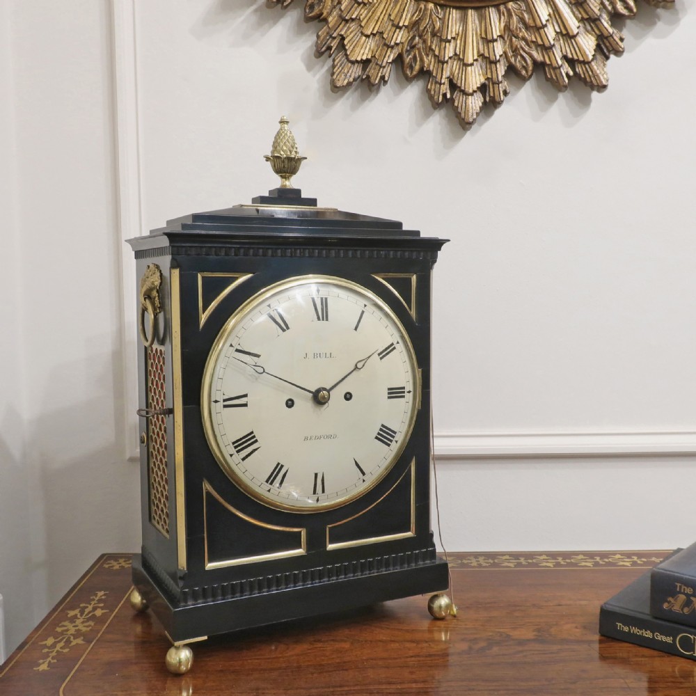 An ebonised William IV bracket clock featuring brass inlays, a pineapple finial, lion mask side handles, ball feet, and a white circular dial with Roman numerals signed J. Bull Bedford.