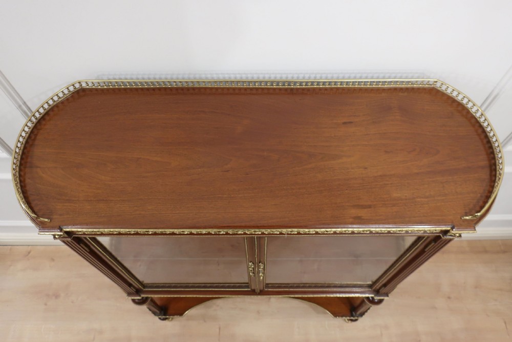 An early twentieth-century French style mahogany display cabinet featuring glazed doors, brass gallery rails, turned legs, and a lower tier. View III