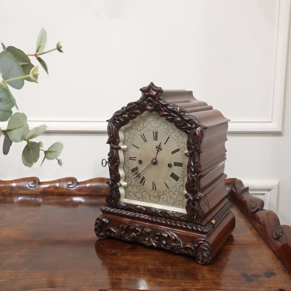 An early Victorian mahogany library clock featuring a heavily carved leafy frame, Roman numeral dial face, and stepped side moulding.