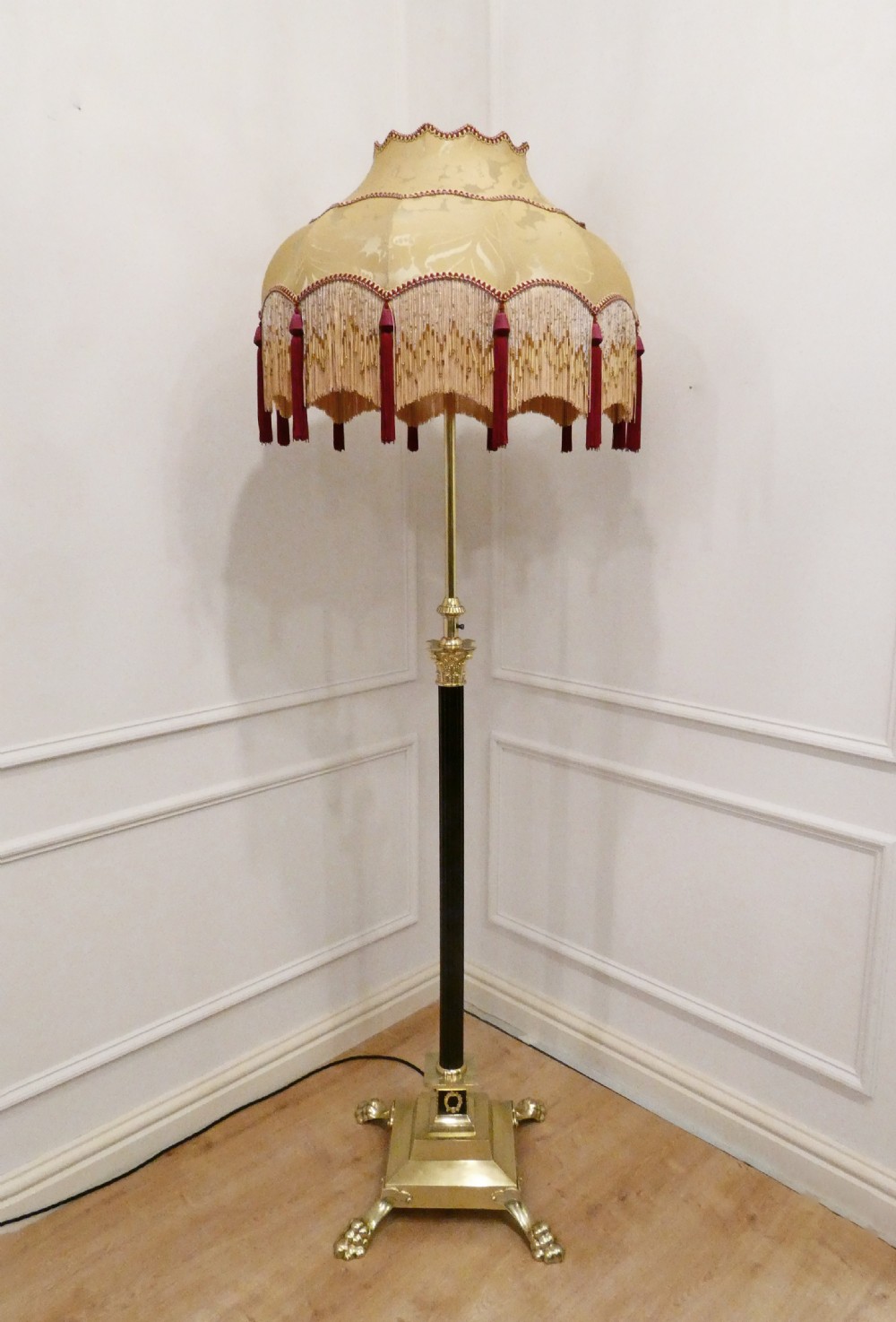 A Victorian brass standard floor lamp featuring a Corinthian capital, stepped base with claw feet, and a fringed cream shade with red tassels.