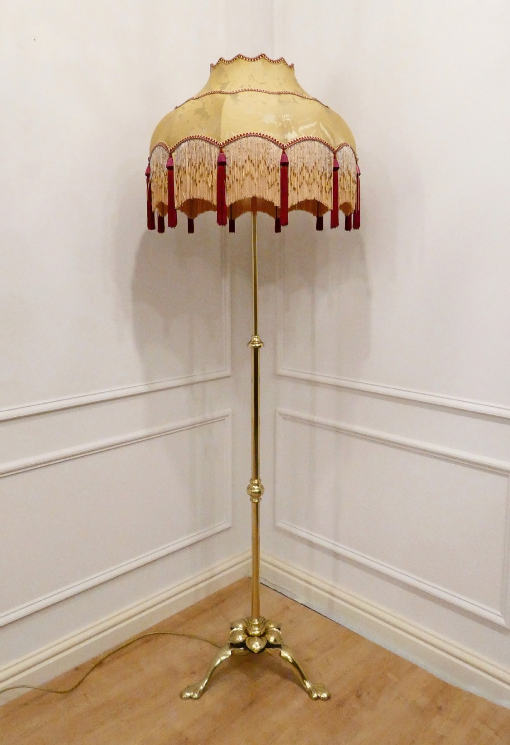 A tall brass standard lamp featuring a tripod base, slender central stem, and a gold fabric dome shade decorated with tassels and fringe.