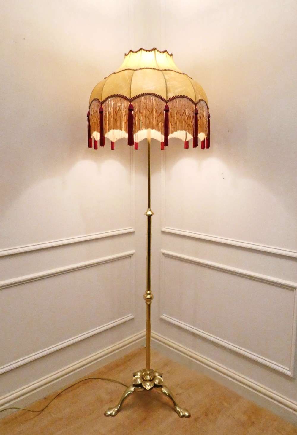 A tall brass standard lamp featuring a tripod base, slender central stem, and a gold fabric dome shade decorated with tassels and fringe. View IX
