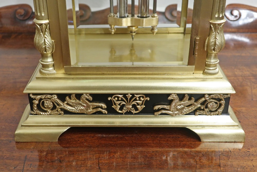 A 19th-century French brass four-glass mantel clock with black decorative corner panels, an enamel dial with Roman numerals, and a visible pendulum movement. View V