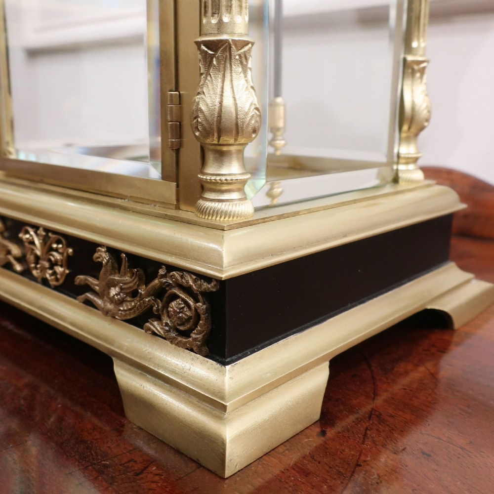 A 19th-century French brass four-glass mantel clock with black decorative corner panels, an enamel dial with Roman numerals, and a visible pendulum movement. View VI
