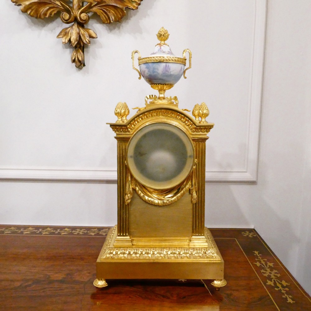 A French Louis XVI style gilt bronze mantel clock featuring a porcelain urn top, hand-painted floral porcelain panels, and Roman numerals on the dial. View XIV