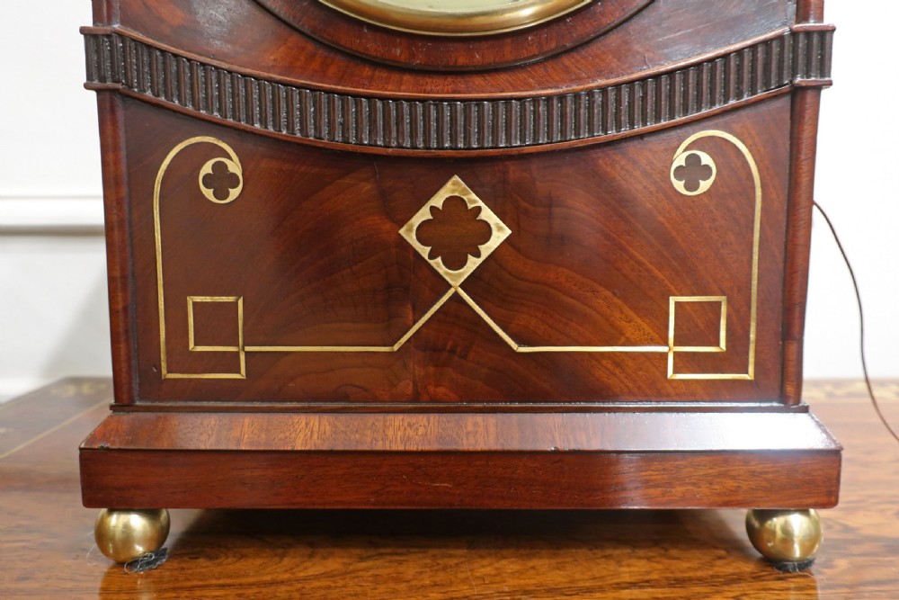 A Georgian mahogany bracket clock with a lancet-arched top, brass geometric inlay, round brass bezel dial, and matching timber wall mounting bracket. View VI