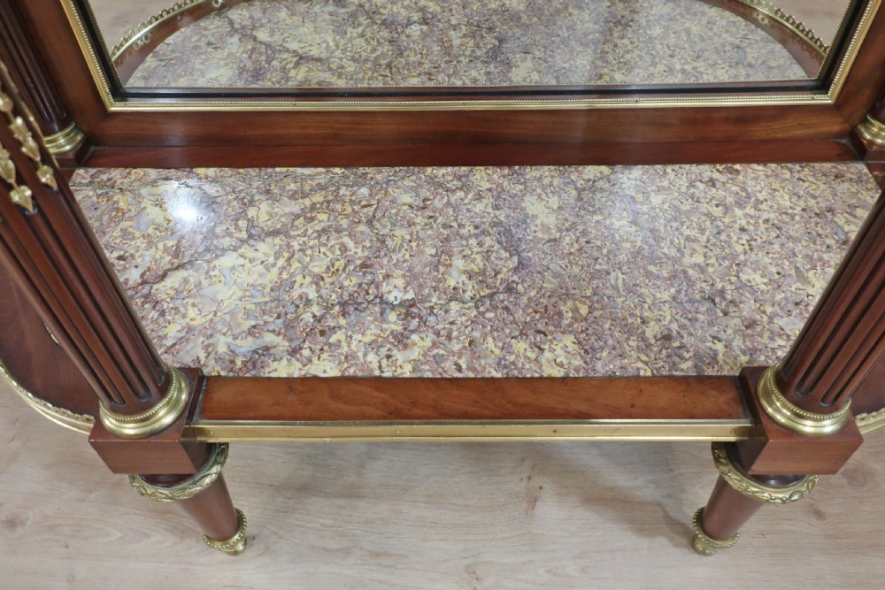 French Louis XVI style mahogany and ormolu-mounted vitrine cabinet featuring curved glass sides, a mirrored back interior, glass shelves, and a marble tier below fluted legs. View XV