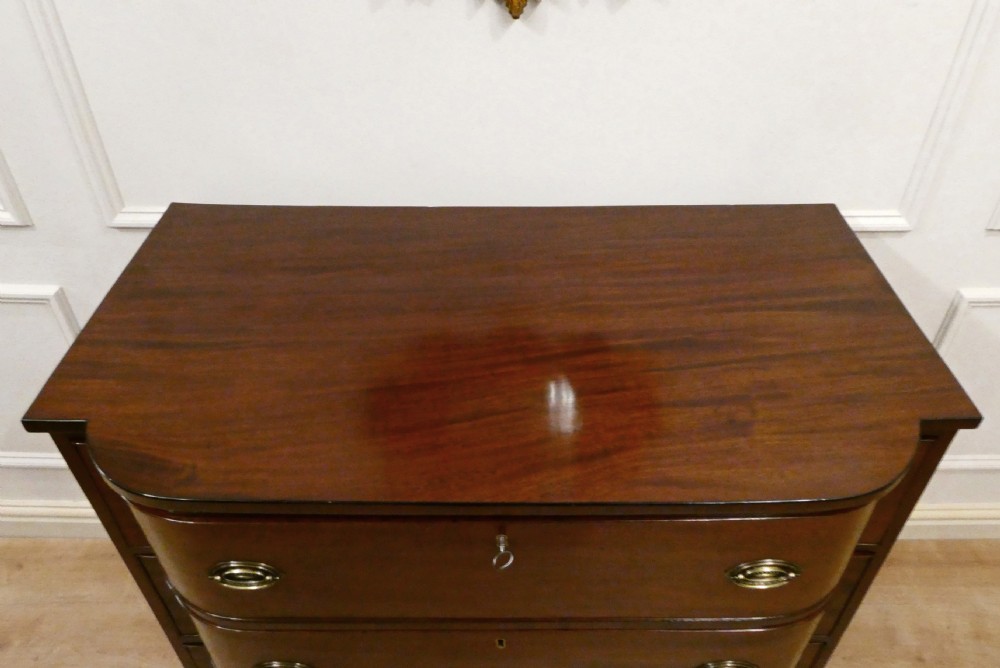 A Regency period mahogany bow-fronted chest of drawers featuring three graduated drawers with brass oval handles and short turned feet. View VI