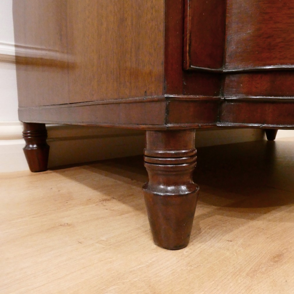 A Regency period mahogany bow-fronted chest of drawers featuring three graduated drawers with brass oval handles and short turned feet. View VIII