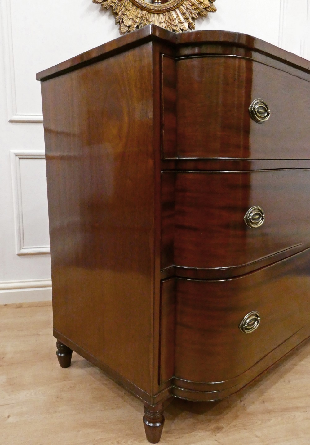 A Regency period mahogany bow-fronted chest of drawers featuring three graduated drawers with brass oval handles and short turned feet. View IX