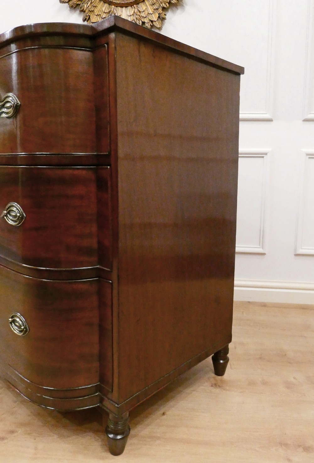 A Regency period mahogany bow-fronted chest of drawers featuring three graduated drawers with brass oval handles and short turned feet. View X