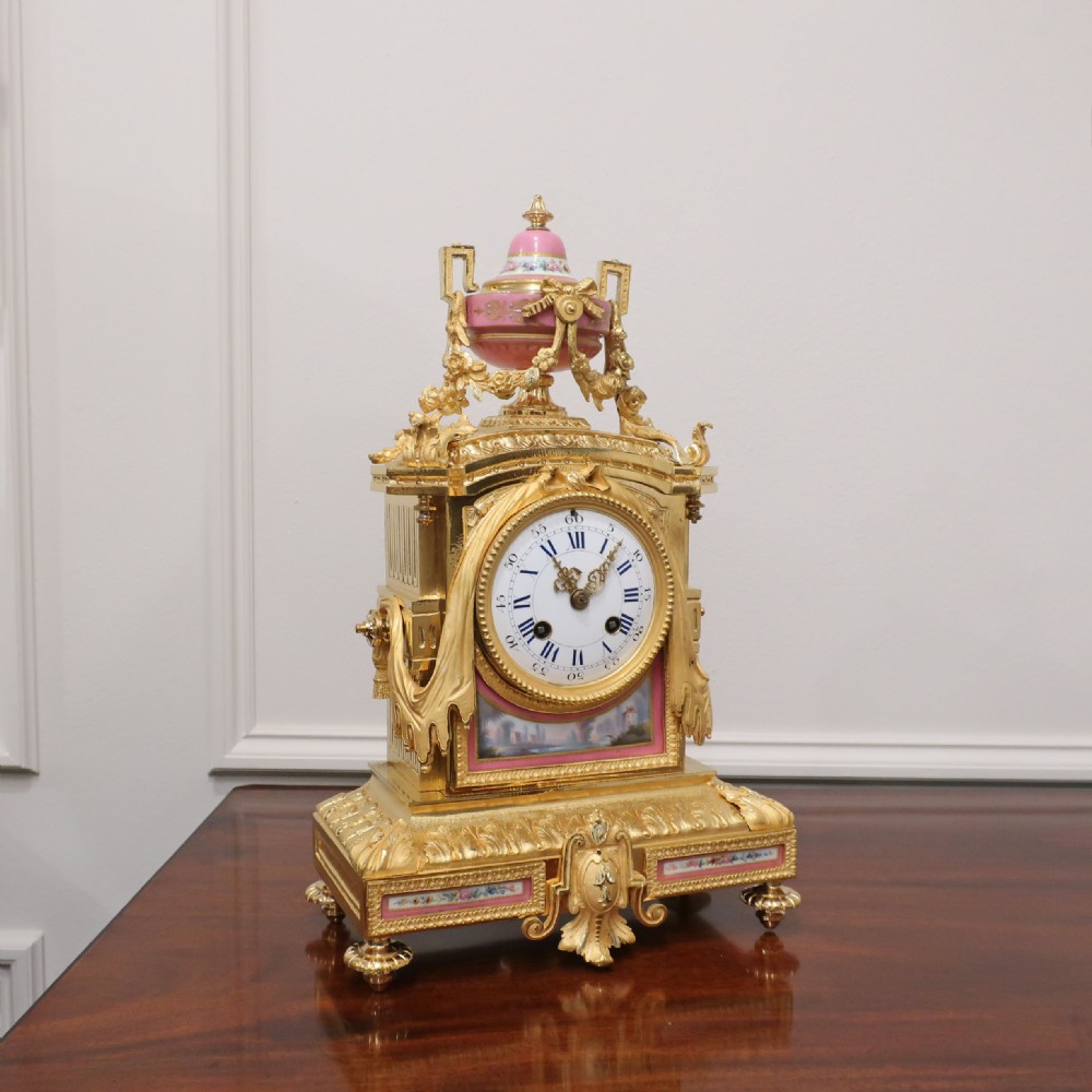 A French gilt bronze mantel clock featuring an urn top finial, pink hand-painted porcelain panels, and a white enamel dial with Roman numerals.