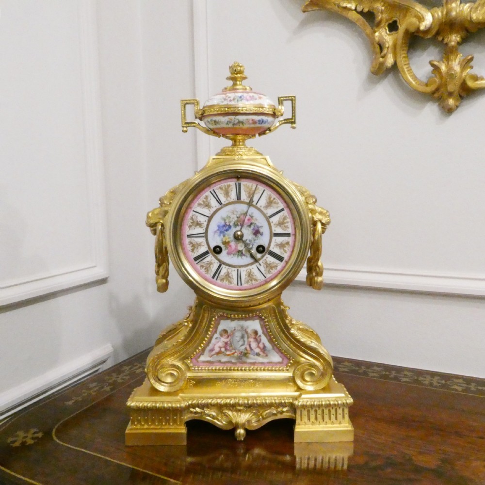 A French Louis XV style gilt bronze mantel clock featuring a hand-painted porcelain dial, lower cherub panel, scroll feet, and an ornamental urn finial.
