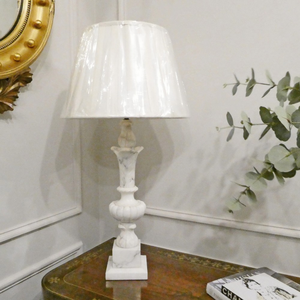 An early 20th-century carved white alabaster table lamp featuring an urn-shaped stem, square plinth base, and a pleated white fabric lampshade.
