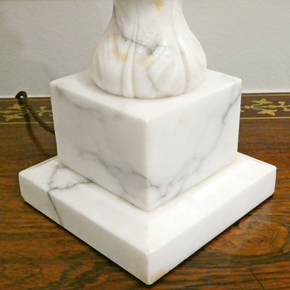 An early 20th-century carved white alabaster table lamp featuring an urn-shaped stem, square plinth base, and a pleated white fabric lampshade. View VII