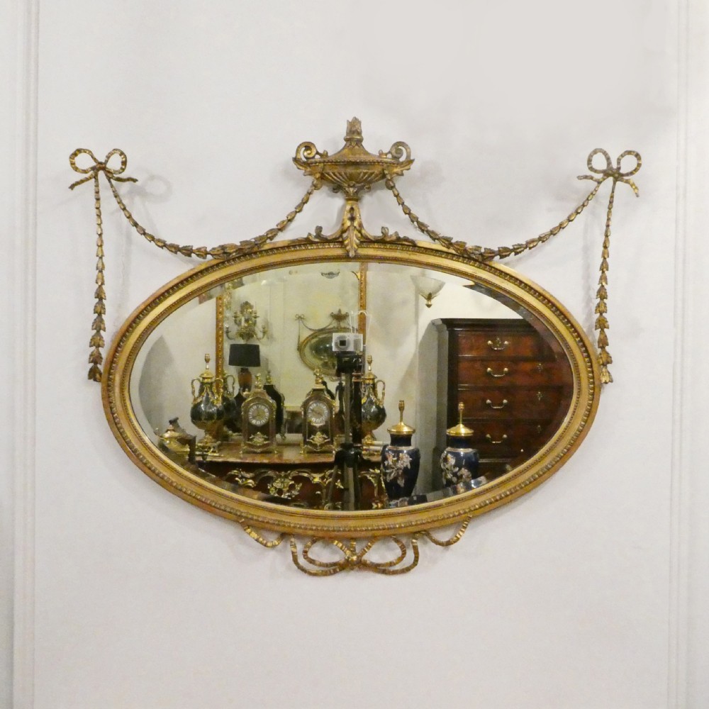An oval giltwood wall mirror featuring Neoclassical urn cresting, suspended husk festoons with bow ribbons, and lower decorative scrollwork.