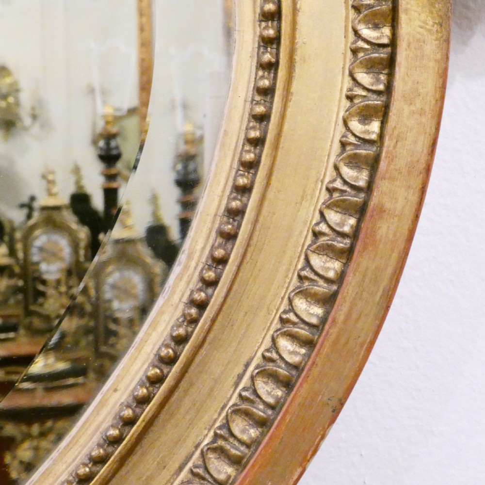An oval giltwood wall mirror featuring Neoclassical urn cresting, suspended husk festoons with bow ribbons, and lower decorative scrollwork. View V