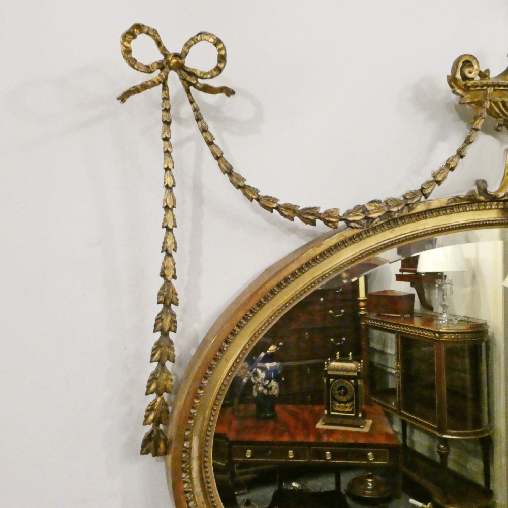 An oval giltwood wall mirror featuring Neoclassical urn cresting, suspended husk festoons with bow ribbons, and lower decorative scrollwork. View VI