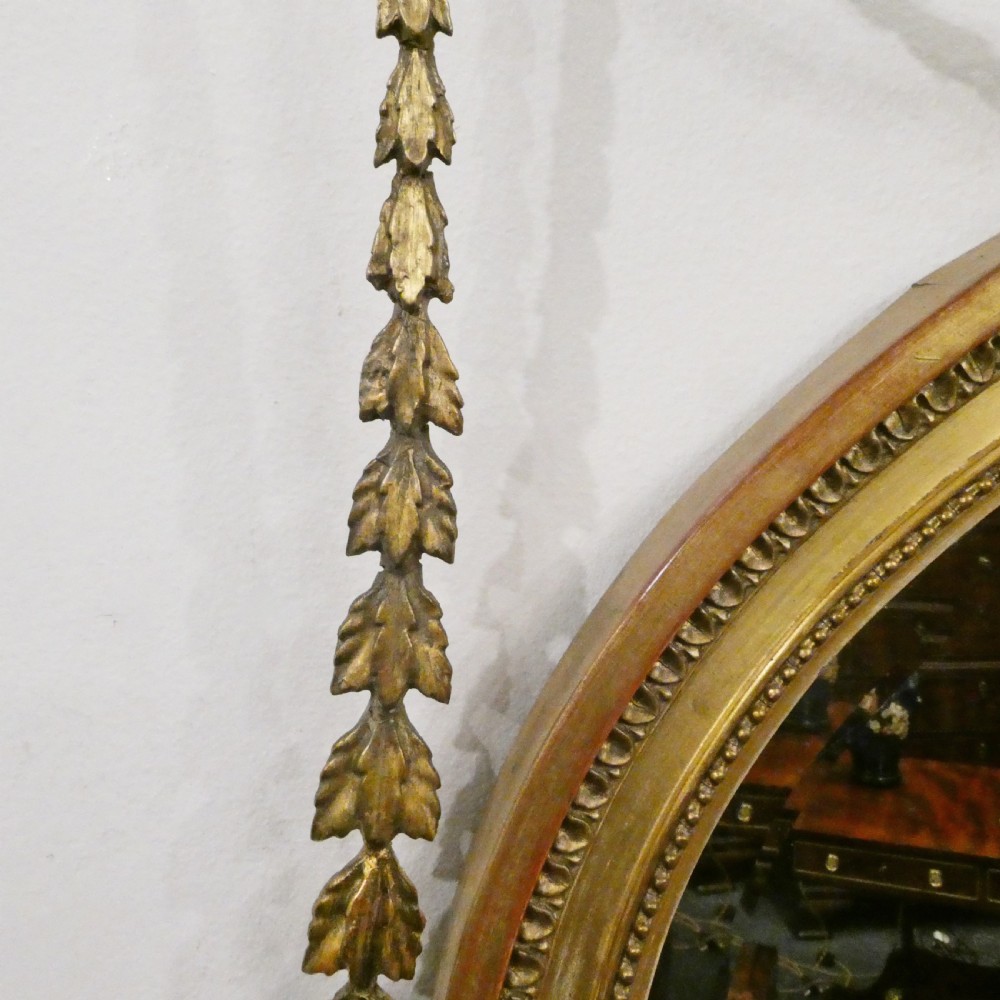 An oval giltwood wall mirror featuring Neoclassical urn cresting, suspended husk festoons with bow ribbons, and lower decorative scrollwork. View VIII