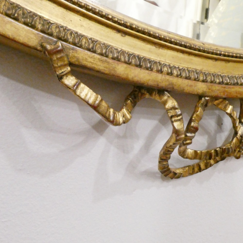 An oval giltwood wall mirror featuring Neoclassical urn cresting, suspended husk festoons with bow ribbons, and lower decorative scrollwork. View X