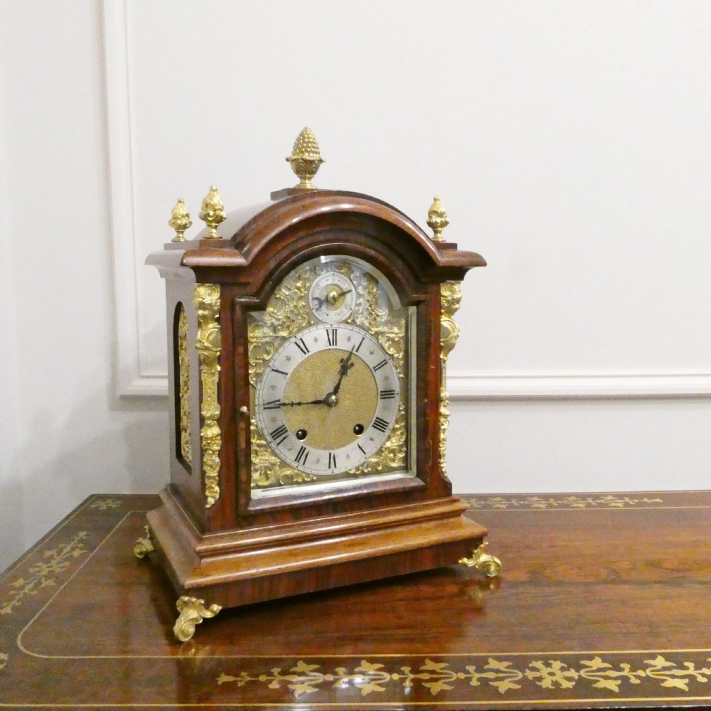 A dark mahogany arched bracket clock featuring gilt brass corner mounts, finials, and a silvered chapter ring dial with brass spandrels.