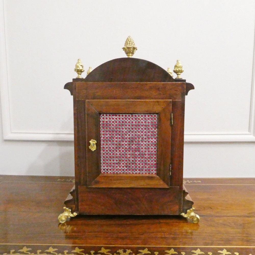 A dark mahogany arched bracket clock featuring gilt brass corner mounts, finials, and a silvered chapter ring dial with brass spandrels. View X
