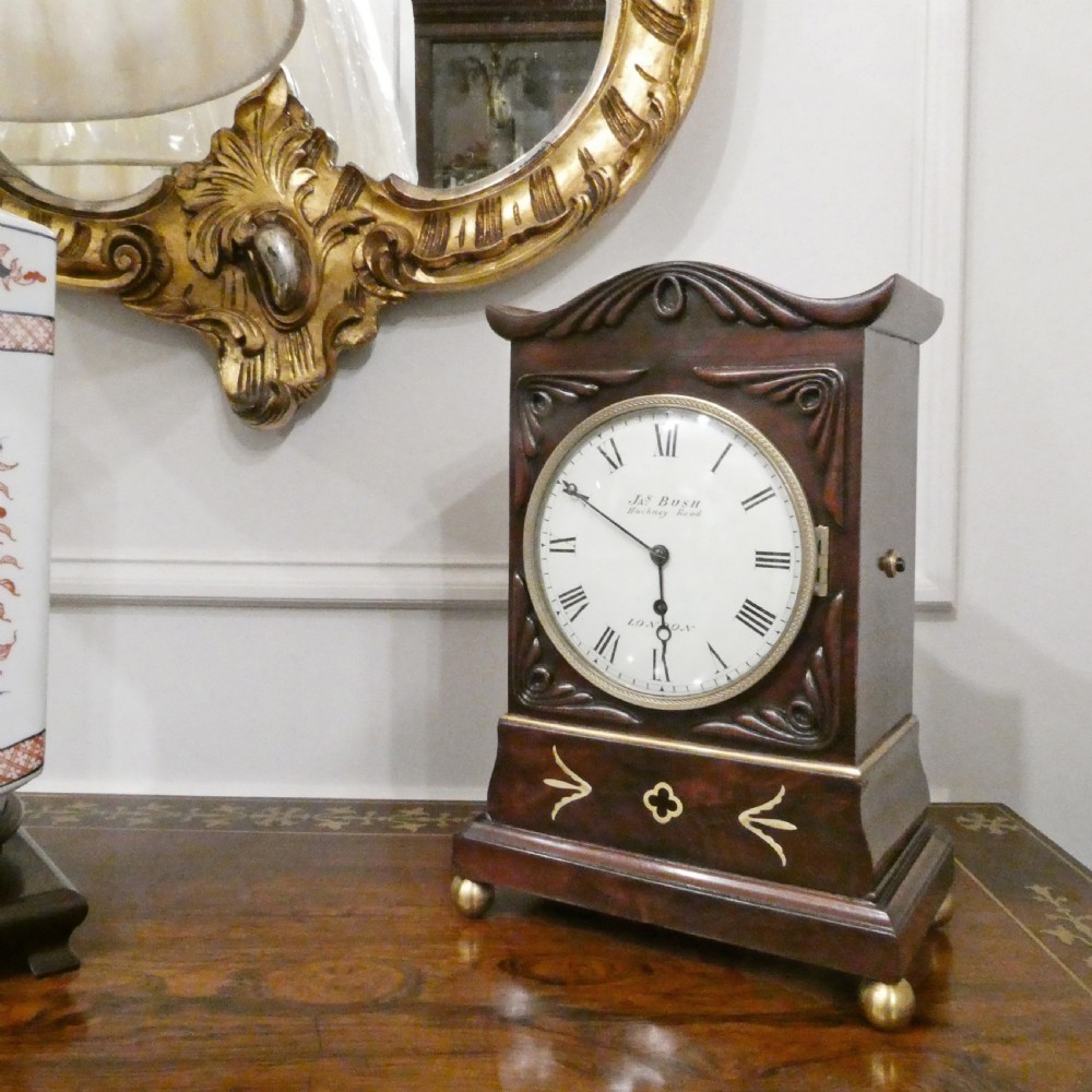 A Georgian mahogany library clock with a carved top crest, brass inlay, white dial with Roman numerals, and brass ball feet.