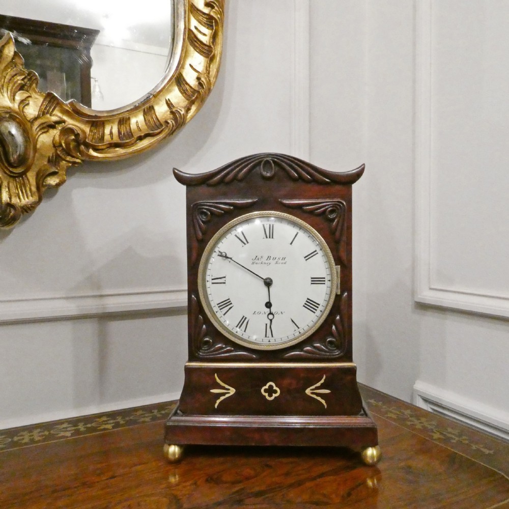 A Georgian mahogany library clock with a carved top crest, brass inlay, white dial with Roman numerals, and brass ball feet. View II