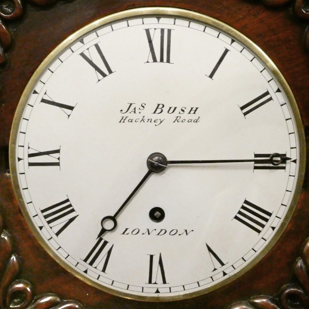 A Georgian mahogany library clock with a carved top crest, brass inlay, white dial with Roman numerals, and brass ball feet. View III