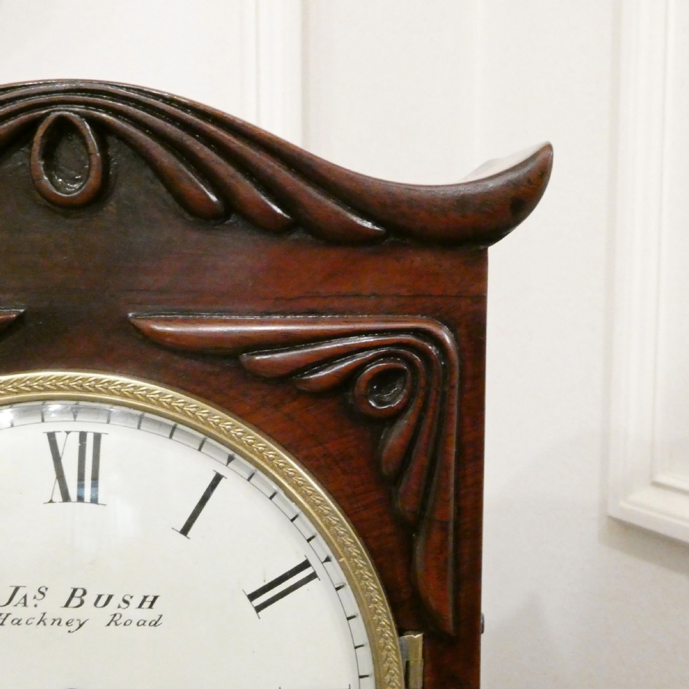 A Georgian mahogany library clock with a carved top crest, brass inlay, white dial with Roman numerals, and brass ball feet. View IV