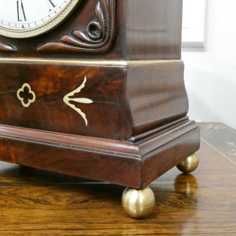 A Georgian mahogany library clock with a carved top crest, brass inlay, white dial with Roman numerals, and brass ball feet. View V
