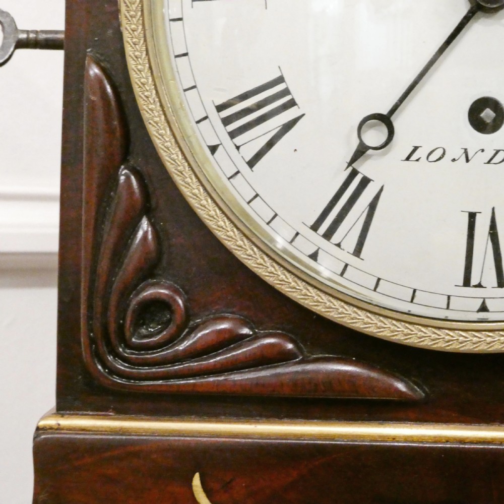 A Georgian mahogany library clock with a carved top crest, brass inlay, white dial with Roman numerals, and brass ball feet. View VI