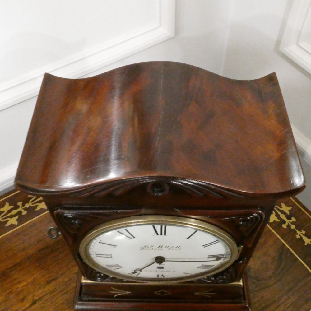 A Georgian mahogany library clock with a carved top crest, brass inlay, white dial with Roman numerals, and brass ball feet. View VII