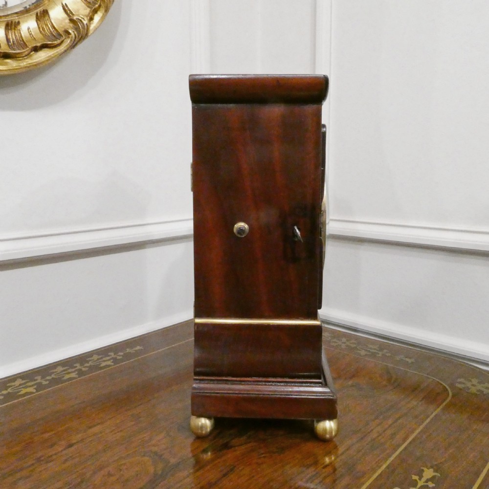 A Georgian mahogany library clock with a carved top crest, brass inlay, white dial with Roman numerals, and brass ball feet. View VIII