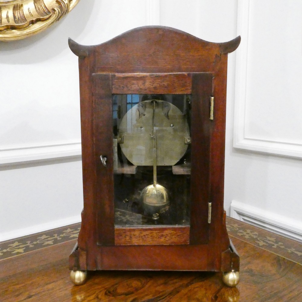 A Georgian mahogany library clock with a carved top crest, brass inlay, white dial with Roman numerals, and brass ball feet. View IX