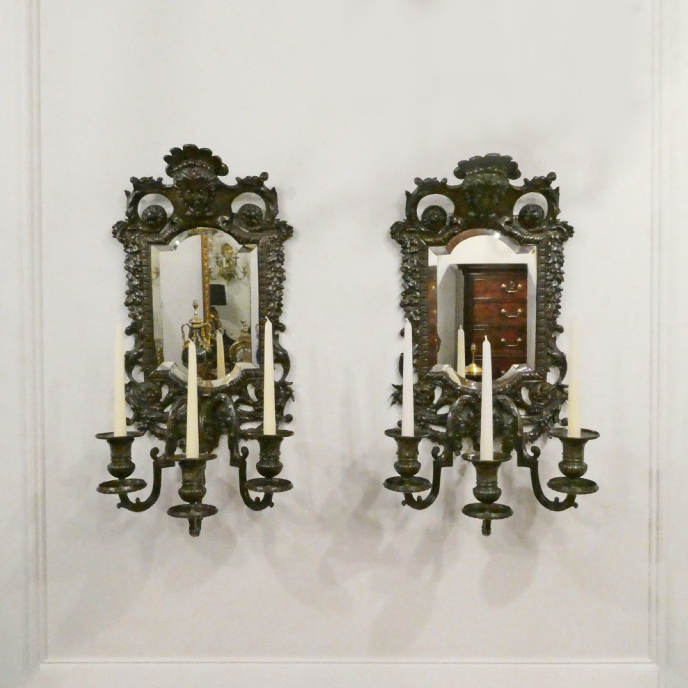 A pair of dark patinated bronze wall girandoles with ornate foliate frames surrounding central mirrored plates and three candle branches each.