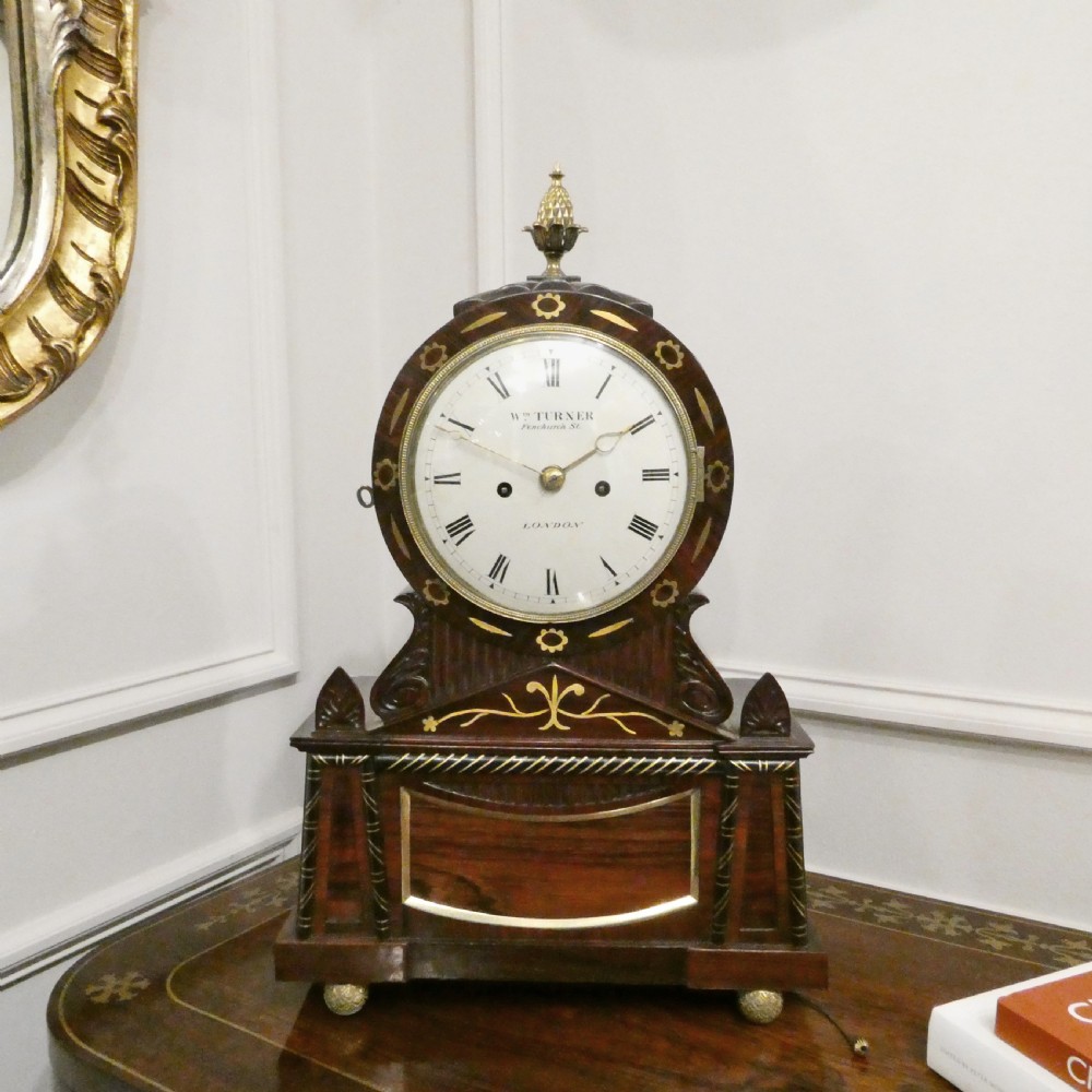 A Georgian rosewood bracket clock featuring intricate brass inlay, a brass pineapple finial, a white circular dial with Roman numerals, and brass ball feet.