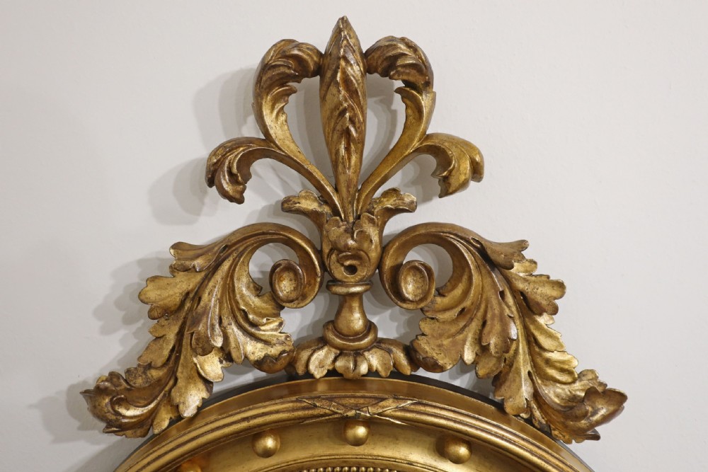 A circular Regency style convex wall mirror with a gilded frame decorated with sphere mouldings, an acanthus leaf cresting, and a foliate bottom apron. View V